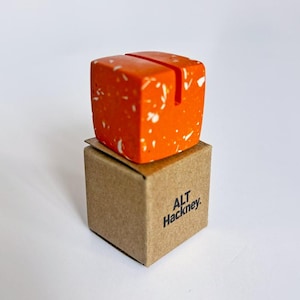 May include: A vibrant orange cube-shaped desk accessory with white flecks, designed to hold notes or photos. It rests on a small, brown cardboard box, with the text "ALT Hackney."