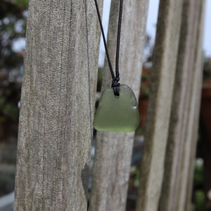 May include: A green sea glass pendant on a black cord necklace. The necklace is hanging from a wooden fence.