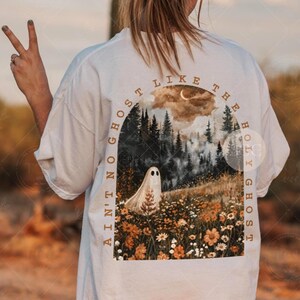 May include: A white t-shirt featuring a graphic on the back. The design shows a ghost in a field of wildflowers and pine trees under a crescent moon. The text "AIN'T NO GHOST LIKE THE HOLY GHOST" is printed in an arch around the illustration.