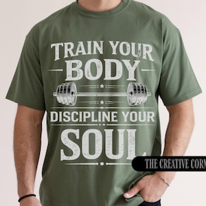 May include: Olive green t-shirt with the text "TRAIN YOUR BODY, DISCIPLINE YOUR SOUL" in white. A barbell graphic is centered between the phrases. The Creative Corner TCC logo is in the bottom right corner.