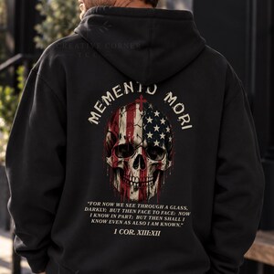 Puede incluir: Una sudadera con capucha negra que presenta un gráfico en la parte posterior de un cráneo humano superpuesto con la bandera estadounidense. Sobre el cráneo está el texto MEMENTO MORI. Debajo del cráneo está el texto FOR NOW WE SEE THROUGH A GLASS, DARKLY; BUT THEN FACE TO FACE: NOW I KNOW IN PART; BUT THEN SHALL I KNOW EVEN AS ALSO I AM KNOWN. 1 COR. XIII:XII.