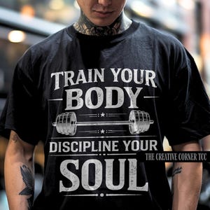 Op de afbeelding: Zwart T-shirt met de witte tekst "TRAIN YOUR BODY - DISCIPLINE YOUR SOUL". Een halterafbeelding is gecentreerd onder "BODY". The Creative Corner TCC staat rechtsonder afgedrukt. De persoon draagt een zwarte pet.