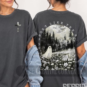 May include: A charcoal gray short-sleeve t-shirt featuring a small cross graphic on the front left chest. The back displays a large, detailed illustration of a ghost in a field of white flowers before a dark forest with the text "NO GHOST LIKE THE H".