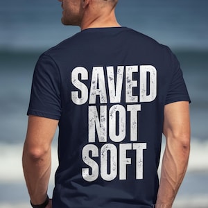 May include: Navy blue t-shirt with the white text "SAVED NOT SOFT" in a distressed font. The shirt is short-sleeved and worn by a person wearing a white baseball cap.