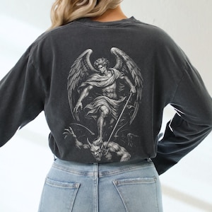 Peut inclure: T-shirt à manches longues gris foncé avec un motif détaillé d'un ange debout sur un démon. L'ange a de grandes ailes et tient un bâton. Le t-shirt est associé à un jean bleu clair.