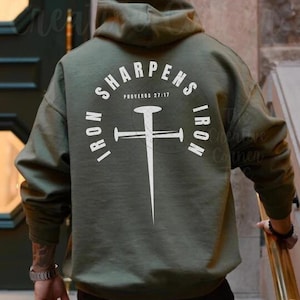 May include: A green hooded sweatshirt with the text "IRON SHARPEN IRON" and "PROVERBS 27:17" printed on the back. The design features a white cross with a nail in the centre.