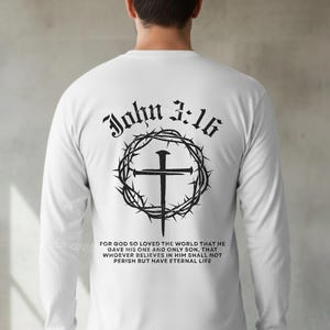 Peut inclure: T-shirt blanc à manches longues avec un motif noir au dos. Le motif comprend le texte "John 3:16" au-dessus d'une croix entourée d'épines. Une citation biblique est en dessous. Le t-shirt porte le texte "THE CREATIVE CORNER TCC" en bas.