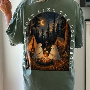 May include: A moss green t-shirt featuring a back graphic of two ghosts roasting marshmallows over a campfire in a forest at night. The scene includes tents, a crescent moon, and the text "AIN'T NO GHOST LIKE THE HOLY GHOST" arched around the image.