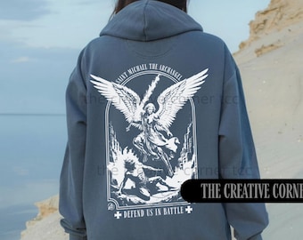 May include: A slate blue hooded sweatshirt featuring a white graphic on the back. The design depicts Saint Michael the Archangel with wings, a sword, and the text "Saint Michael the Archangel" and "Defend Us In Battle" with cross symbols.