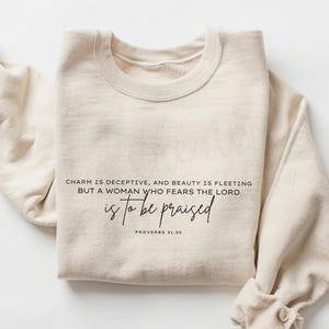 May include: A cream-coloured sweatshirt featuring the text "CHARM IS DECEPTIVE, AND BEAUTY IS FLEETING BUT A WOMAN WHO FEARS THE LORD is to be praised PROVERBS 31:30" in black script. The sweatshirt has a crew neck and long sleeves.