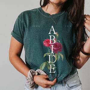 May include: A green t-shirt with the word "ABIDE" printed vertically in white letters. The letters are surrounded by pink and red roses. The text "JOHN 15:4" is printed in white at the top right corner of the shirt.