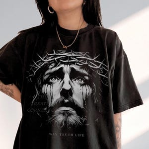 Może przedstawiać: Czarna koszulka z szarą grafiką mężczyzny w koronie cierniowej, ze łzami. Pod obrazem wydrukowano tekst „WAY TRUTH LIFE”. Koszulka ma krótkie rękawy i luźny krój. Obraz zawiera również tekst „THE CREATION CORNER”.