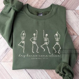 May include: An olive green sweatshirt featuring a white graphic of four dancing skeletons. The text "dry bones come alive" and "EZEKIEL 37:5" are printed below the skeletons. The sweatshirt is folded on a light wood surface.