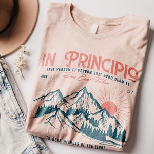May include: A light pink t-shirt with a mountain and sun graphic. The shirt features the text "IN PRINCIPIO" in red, along with other text in red and black. The shirt is folded and placed on a white surface.