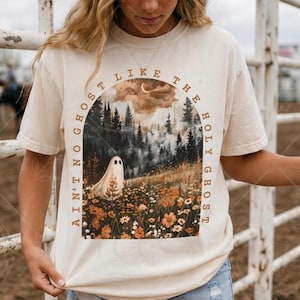 May include: A cream-colored short-sleeve t-shirt featuring a graphic print of a ghost in a field of wildflowers and pine trees under a crescent moon. The text "AIN'T NO GHOST LIKE THE HOLY GHOST" arches over the illustration in a brown font.
