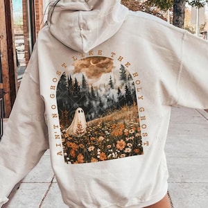Peut inclure: Un sweat à capuche couleur sable avec un motif au dos représentant un fantôme dans un champ de fleurs sauvages et de pins sous un ciel lunaire. Le texte entourant le graphique arqué indique : "AIN'T NO GHOST LIKE THE HOLY GHOST."