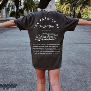 May include: A dark gray t-shirt with white text and a graphic of sheep. The text reads "The Parable of The Lost Sheep" and includes a Bible verse reference.