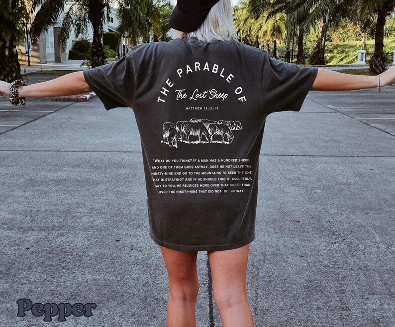 May include: A dark gray t-shirt with white text and a graphic of sheep. The text reads "The Parable of The Lost Sheep" and includes a Bible verse reference.