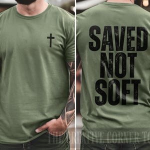 May include: Olive green t-shirt with a black cross on the front and the words "SAVED NOT SOFT" printed in black on the back. The shirt is a crew neck style.
