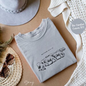 May include: A gray t-shirt with a black and white illustration of sheep and the text "Matthew 18:11-13". The t-shirt is on a brown surface with a gray hat, a white crocheted cardigan, and a fan.