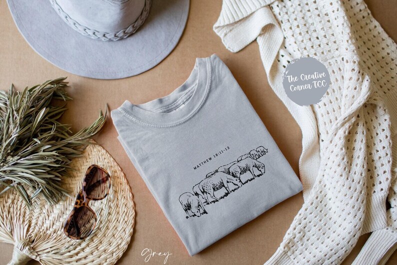 May include: A gray t-shirt with a black and white illustration of sheep and the text "Matthew 18:11-13". The t-shirt is on a brown surface with a gray hat, a white crocheted cardigan, and a fan.