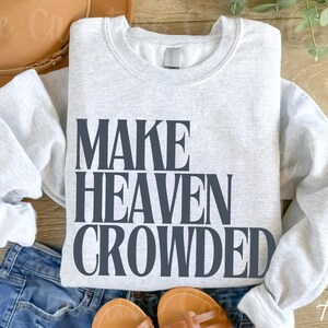 May include: A light grey sweatshirt featuring the text "MAKE HEAVEN CROWDED" in dark grey. The sweatshirt is placed on blue jeans, with a brown hat and sandals in the background. The word "Ash" is in the bottom right corner.