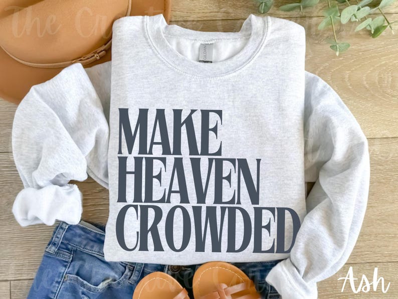 May include: A light grey sweatshirt featuring the text "MAKE HEAVEN CROWDED" in dark grey. The sweatshirt is placed on blue jeans, with a brown hat and sandals in the background. The word "Ash" is in the bottom right corner.