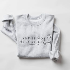 May include: A light gray sweatshirt with the text "AND IF NOT HE IS STILL Good" and "DANIEL 3:18" printed in black. A floral design is next to the word "Good". The sleeves are cuffed.