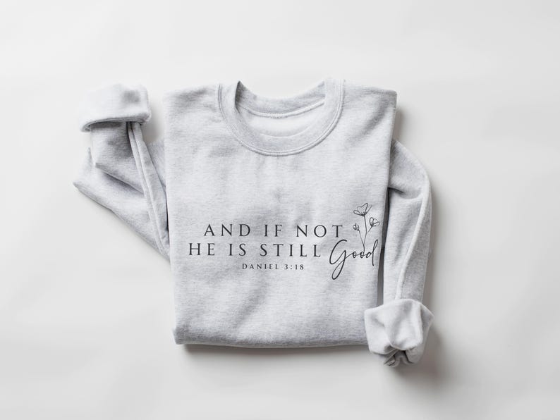 May include: A light gray sweatshirt with the text "AND IF NOT HE IS STILL Good" and "DANIEL 3:18" printed in black. A floral design is next to the word "Good". The sleeves are cuffed.