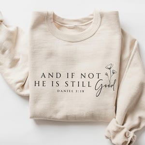 May include: A cream-colored sweatshirt with the text "AND IF NOT HE IS STILL Good" and "DANIEL 3:18" printed in black. A floral design is next to the word "Good". The sleeves are cuffed.