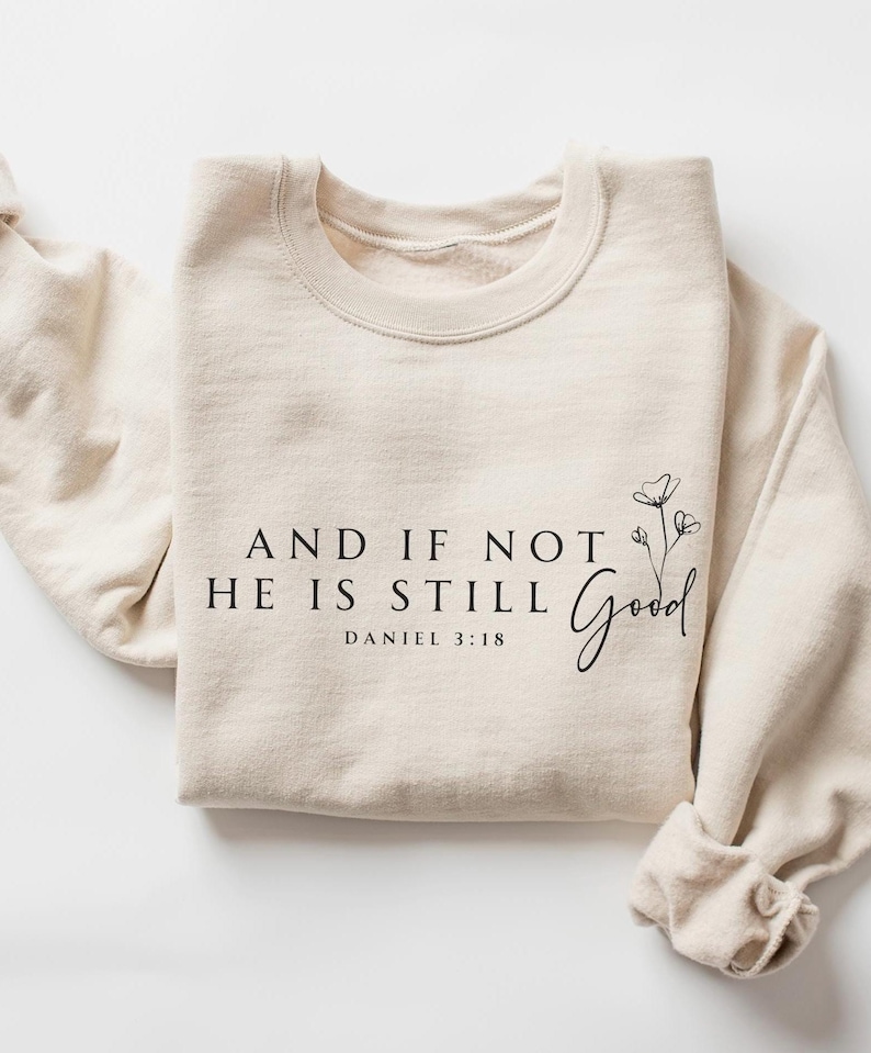 May include: A cream-colored sweatshirt with the text "AND IF NOT HE IS STILL Good" and "DANIEL 3:18" printed in black. A floral design is next to the word "Good". The sleeves are cuffed.