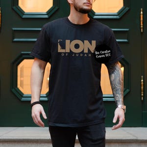 K&ouml;nnte beinhalten: Schwarzes T-Shirt mit einem goldenen Grafikdesign, auf dem "Lion of Judah" und "The Creative Corner TCC" steht.