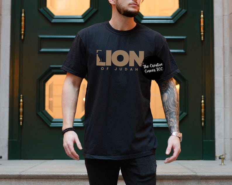 K&ouml;nnte beinhalten: Schwarzes T-Shirt mit einem goldenen Grafikdesign, auf dem "Lion of Judah" und "The Creative Corner TCC" steht.