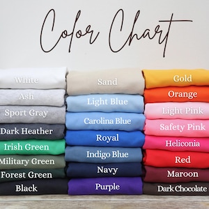 May include: A color chart displays folded fabric samples in various hues. The chart includes colors like white, ash, sport gray, dark heather, and black. Other colors include sand, light blue, royal, navy, and purple. The text "Color Chart" is written in cursive.