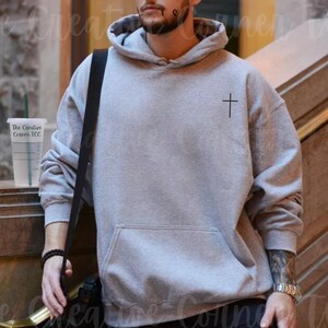 May include: A gray hooded sweatshirt with a small black cross embroidered on the chest. The sweatshirt has a drawstring hood and a kangaroo pocket.