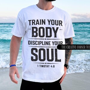 Op de afbeelding: Wit T-shirt met de zwarte tekst "TRAIN YOUR BODY, DISCIPLINE YOUR SOUL" en "1 TIMOTHY 4:8" eronder. Het T-shirt heeft een klassieke pasvorm.