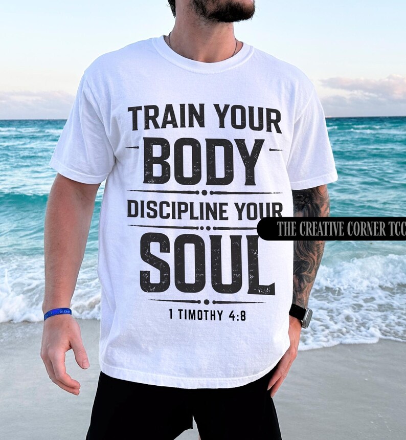 Op de afbeelding: Wit T-shirt met de zwarte tekst "TRAIN YOUR BODY, DISCIPLINE YOUR SOUL" en "1 TIMOTHY 4:8" eronder. Het T-shirt heeft een klassieke pasvorm.