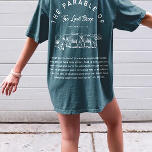 May include: Teal blue t-shirt with white text and a graphic of sheep. The text reads "The Parable of The Lost Sheep Matthew 18:11-13" and includes a passage from the Bible.