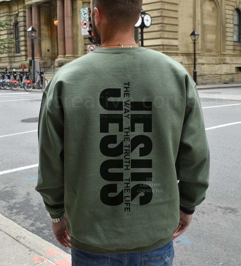 May include: Olive green sweatshirt with the word "JESUS" printed vertically in large black letters. The text "THE WAY THE TRUTH THE LIFE" is printed vertically next to the word "JESUS". The Creative Corner TCC is printed at the bottom.