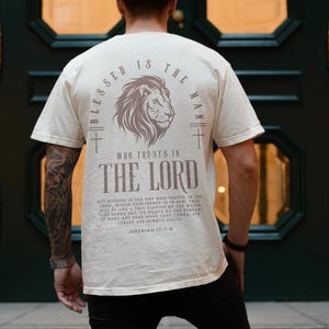 Puede incluir: Camiseta color crema con un gr&aacute;fico de le&oacute;n y el texto "Blessed is the Man Who Trusts in the Lord". La parte posterior de la camiseta tambi&eacute;n presenta un dise&ntilde;o de cruz y un vers&iacute;culo b&iacute;blico. La camiseta est&aacute; hecha de un material suave.