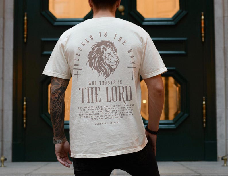 Puede incluir: Camiseta color crema con un gr&aacute;fico de le&oacute;n y el texto "Blessed is the Man Who Trusts in the Lord". La parte posterior de la camiseta tambi&eacute;n presenta un dise&ntilde;o de cruz y un vers&iacute;culo b&iacute;blico. La camiseta est&aacute; hecha de un material suave.