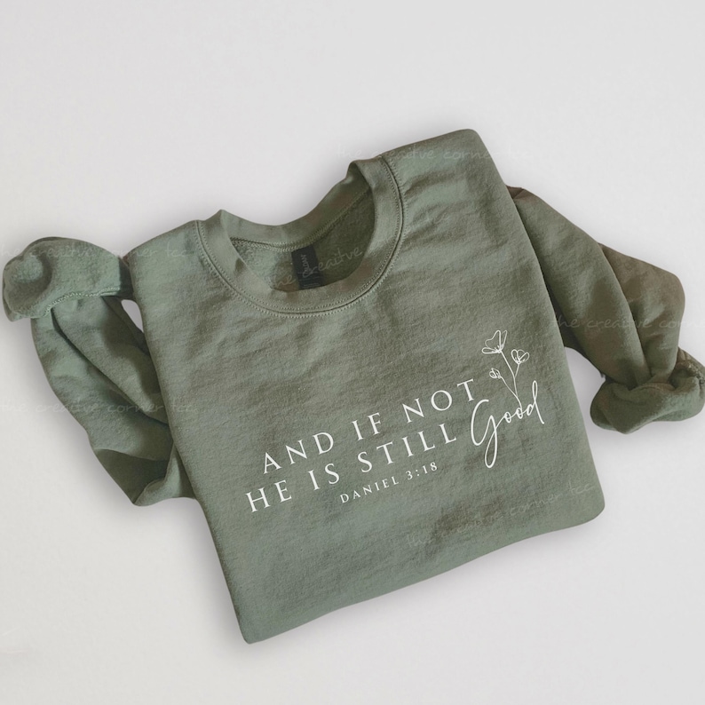 May include: Sage green sweatshirt with the text "AND IF NOT HE IS STILL Good" and "DANIEL 3:18" in white. A delicate floral design is next to the word "Good". The long-sleeved top is made of a soft material.