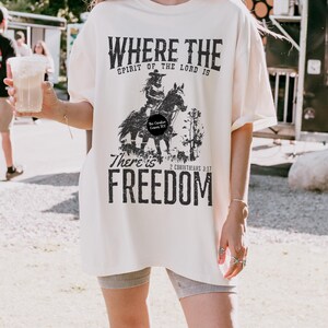 May include: Cream-colored t-shirt with a distressed black graphic of a person on a horse, with the words "WHERE THE SPIRIT OF THE LORD IS THERE IS FREEDOM" and the Bible verse 2 Corinthians 3:17. The shirt is oversized.