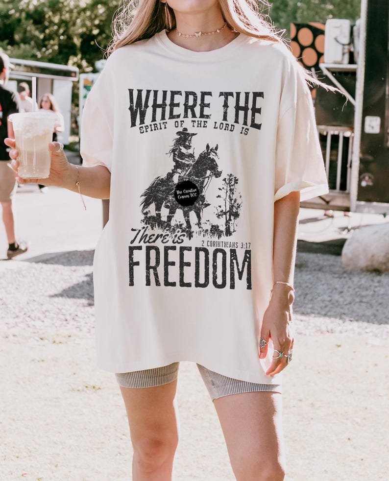 May include: Cream-colored t-shirt with a distressed black graphic of a person on a horse, with the words "WHERE THE SPIRIT OF THE LORD IS THERE IS FREEDOM" and the Bible verse 2 Corinthians 3:17. The shirt is oversized.