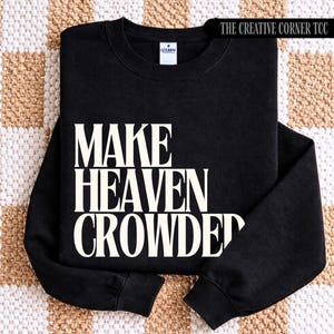 May include: Black crew neck sweatshirt featuring the white text "MAKE HEAVEN CROWDED". The phrase "THE CREATIVE CORNER TCC" is printed in white on the top right corner. The sweatshirt is folded on a patterned surface.