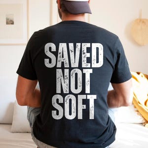May include: Black t-shirt with the white text "SAVED NOT SOFT" printed on the back. The shirt is worn by a person sitting on a bed. The text has a distressed, textured appearance.