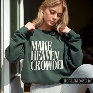 May include: A dark green crew neck sweatshirt featuring the white text "MAKE HEAVEN CROWDED". The long-sleeved top has a relaxed fit. The Creative Corner TCC logo is visible in the bottom right corner.