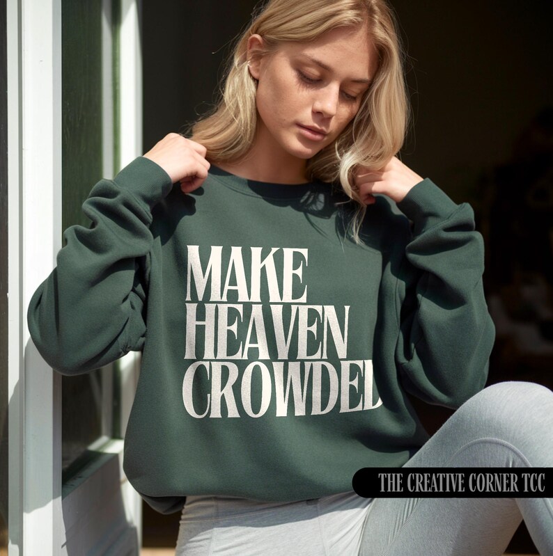May include: A dark green crew neck sweatshirt featuring the white text "MAKE HEAVEN CROWDED". The long-sleeved top has a relaxed fit. The Creative Corner TCC logo is visible in the bottom right corner.