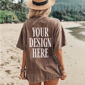 Könnte beinhalten: Braunes T-Shirt mit dem weißen Text "YOUR DESIGN HERE". Das Model trägt einen Strohhut mit schwarzem Band. Das Shirt hat eine lockere Passform, ideal für individuelle Designs.