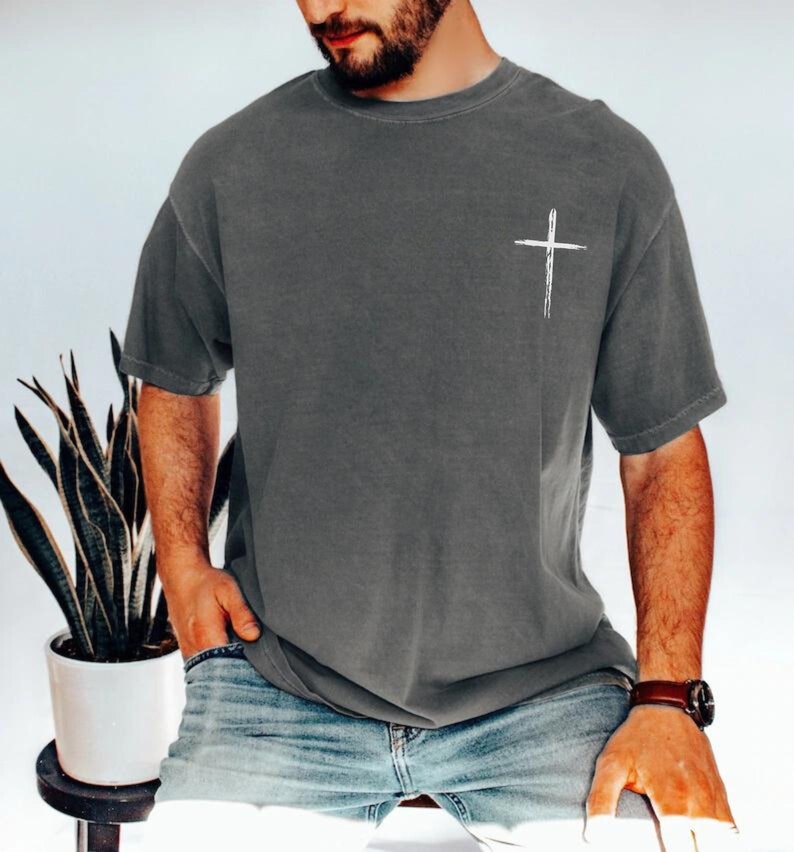 May include: Dark gray t-shirt with a white cross graphic on the left chest. The shirt has short sleeves and a relaxed fit. The model is wearing blue jeans and a brown watch.